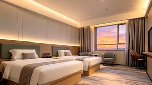 A hotel room with two beds, a desk, a chair, and a large window with a sunset view. - Wanlin Yijing Holiday Hotel (Foshan)