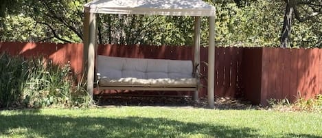 A covered outdoor seating area with a cushioned bench, surrounded by a wooden fence and greenery.