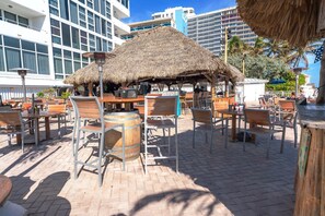 Zona pranzo all'aperto con tavoli e sedie in legno, una struttura con tetto di paglia e vista su edifici moderni sullo sfondo.