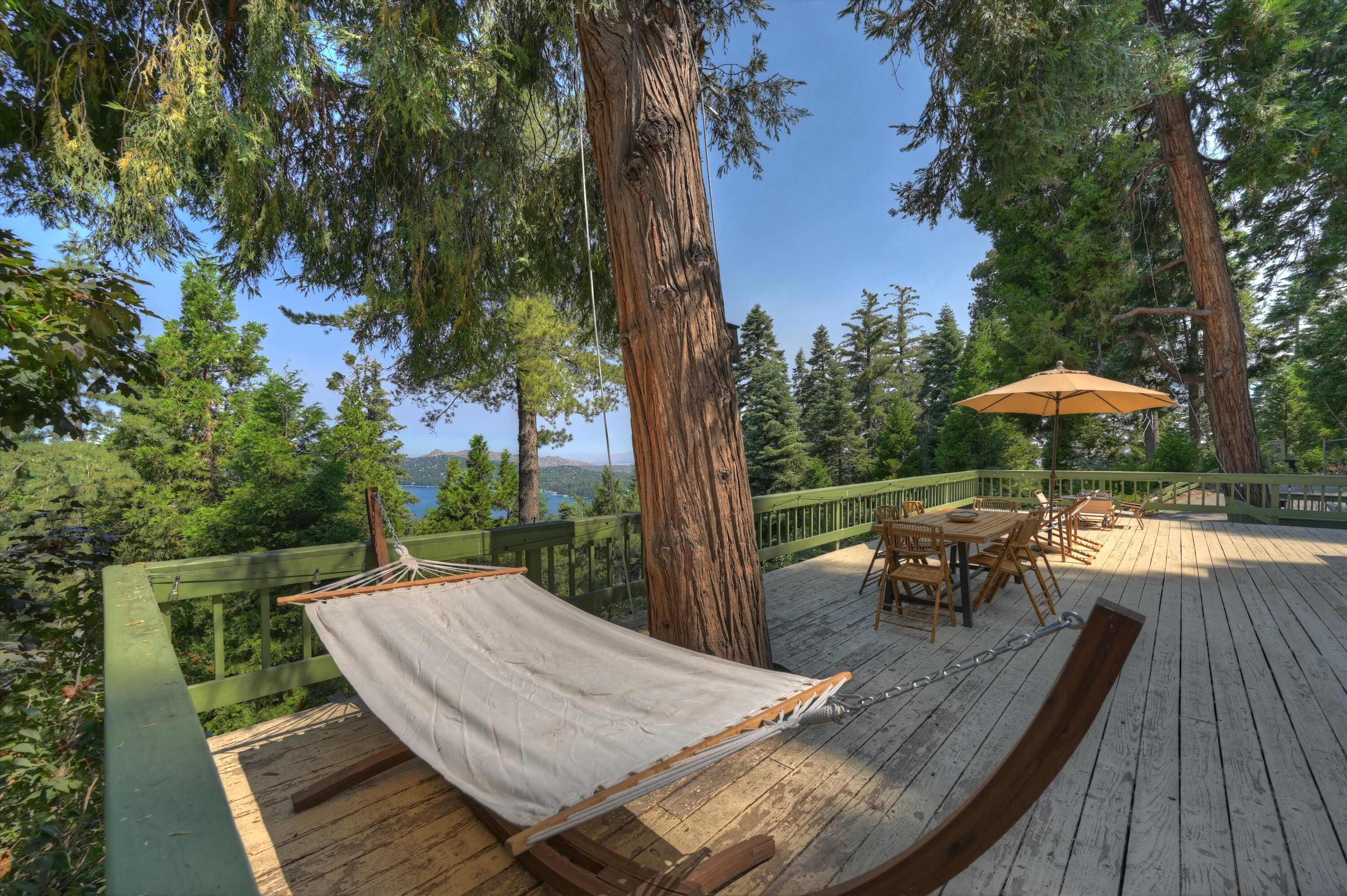 Um deck de madeira com uma rede, móveis de exterior e vista para um lago rodeado por árvores.