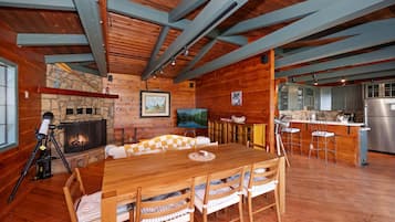 A cozy cabin interior with a stone fireplace, wooden dining table, and kitchen area.