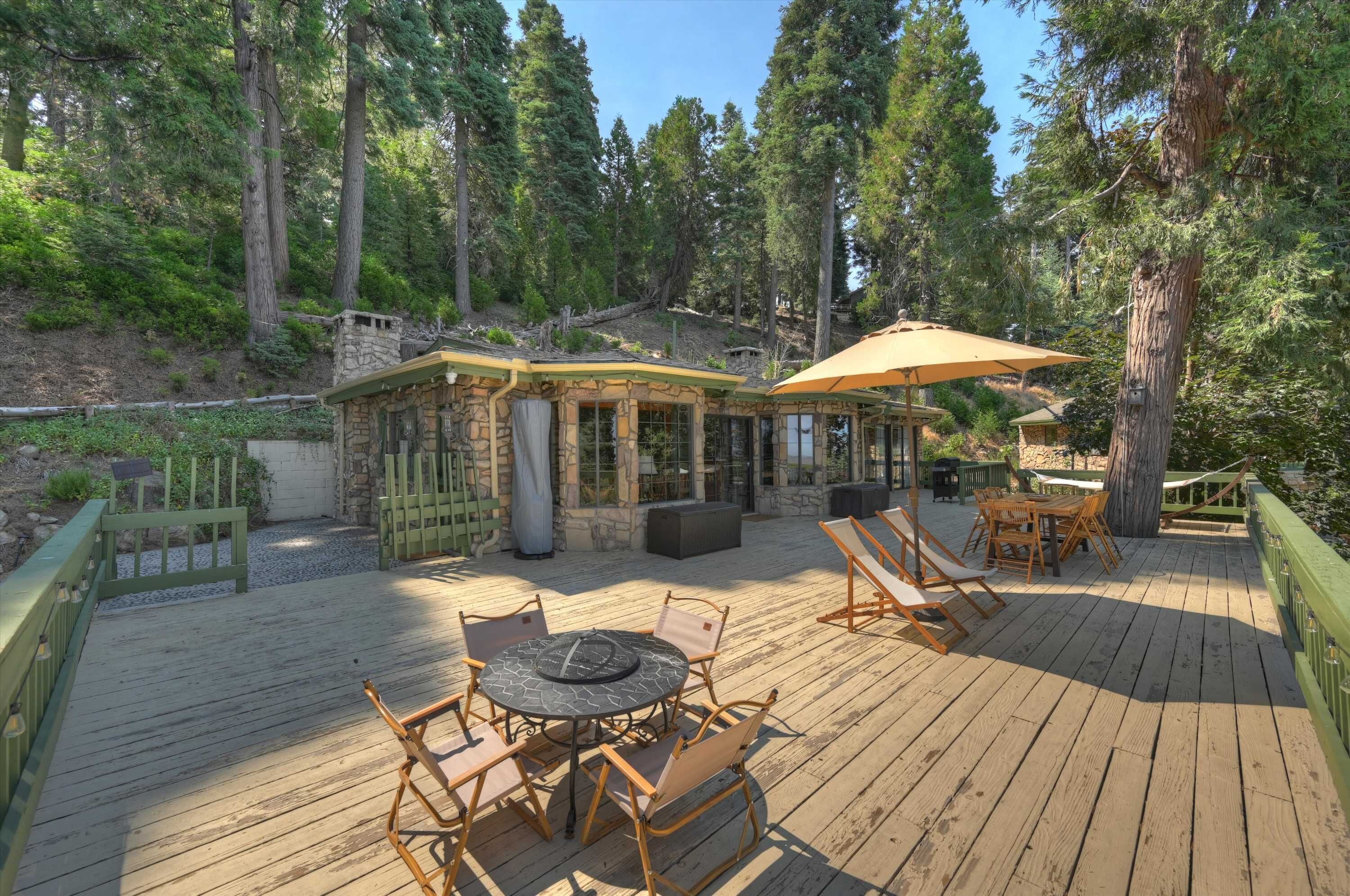 A wooden deck with outdoor furniture, a stone building, and a forested hillside.
