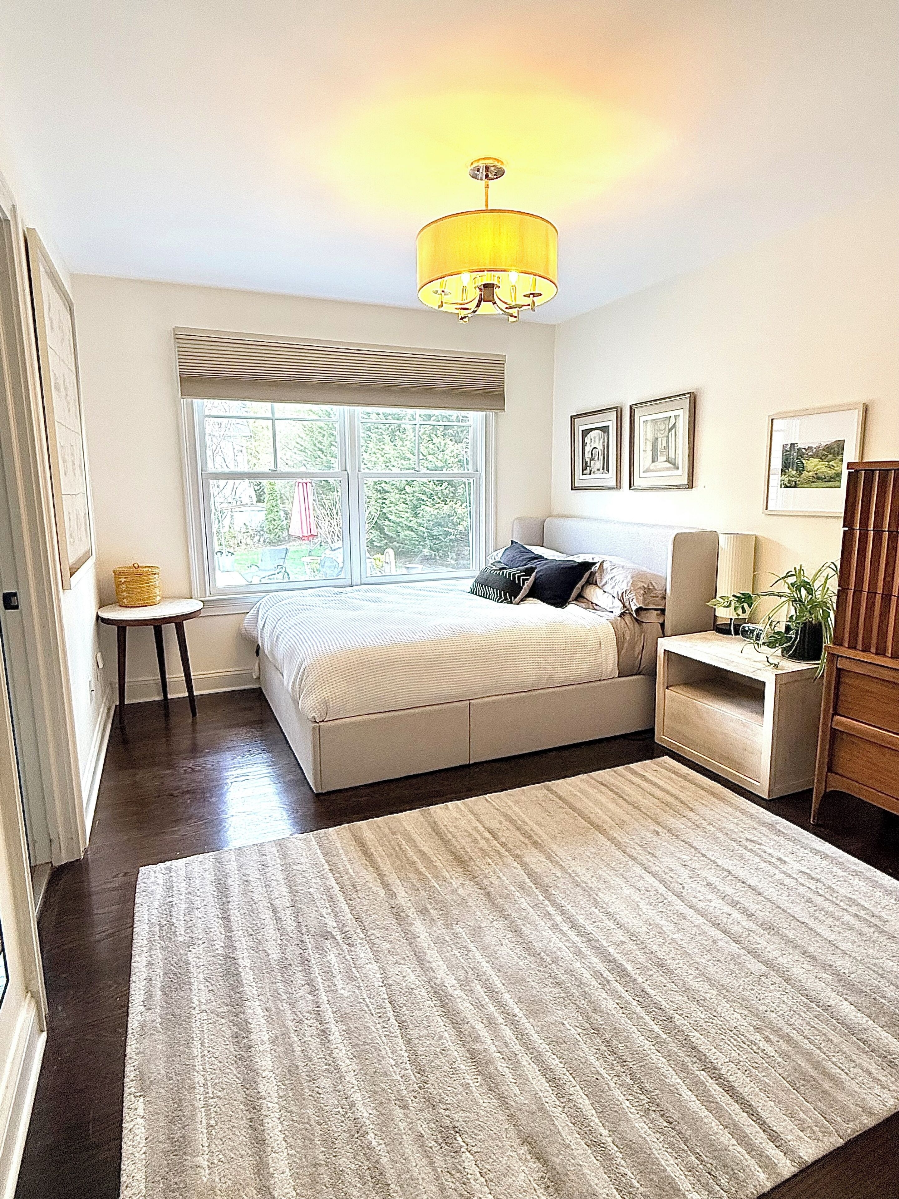 A bedroom with a bed, bedside table, lamp, and a window with a view of the outdoors.
