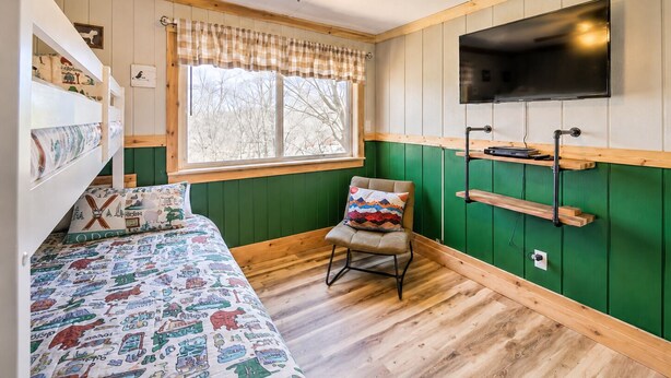 A room with a bunk bed, a chair, a television mounted on the wall, and a window with a view of trees. - Charming 3-bedroom condo with Pool, Hot Tub, AC  & Games (Beech Mountain)