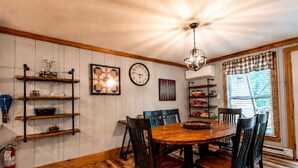 A dining area with a round wooden table, six chairs, a chandelier, wall-mounted shelves, a clock, and a window with blinds. - Charming 3-bedroom condo with Pool, Hot Tub, AC  & Games (Beech Mountain)