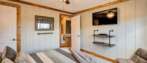 A bedroom with a bed, a ceiling fan, a TV mounted on the wall, and a door leading to another room.