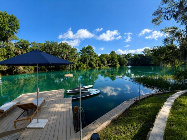 A wooden deck with lounge chairs and a blue umbrella, overlooking a calm lake with paddle boats.