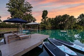 A wooden dock with lounge chairs and a covered seating area, a calm body of water, and a row of paddle boats.