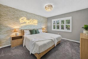 Un dormitorio con cama, mesitas de noche, un letrero de neón en la pared, una ventana con persianas y una lámpara en el techo.