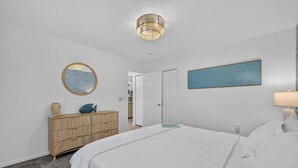 Un dormitorio con cama, cómoda, espejo, un cuadro y una puerta que da a otra habitación.