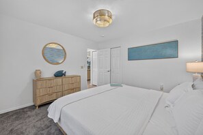 A bedroom with a bed, a dresser, a mirror, a painting, and a door leading to another room.
