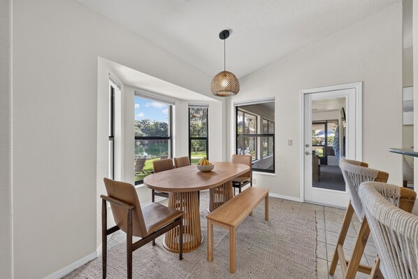 A dining area with a round table and chairs, a bench, and a view of a garden through large windows.