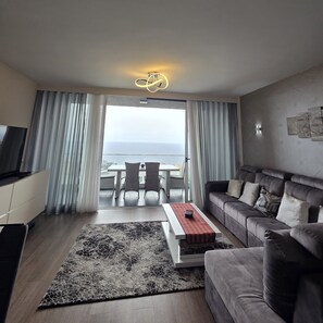 Un salon moderne avec un canapé, une table basse et un téléviseur. Il y a un balcon avec vue sur la mer.