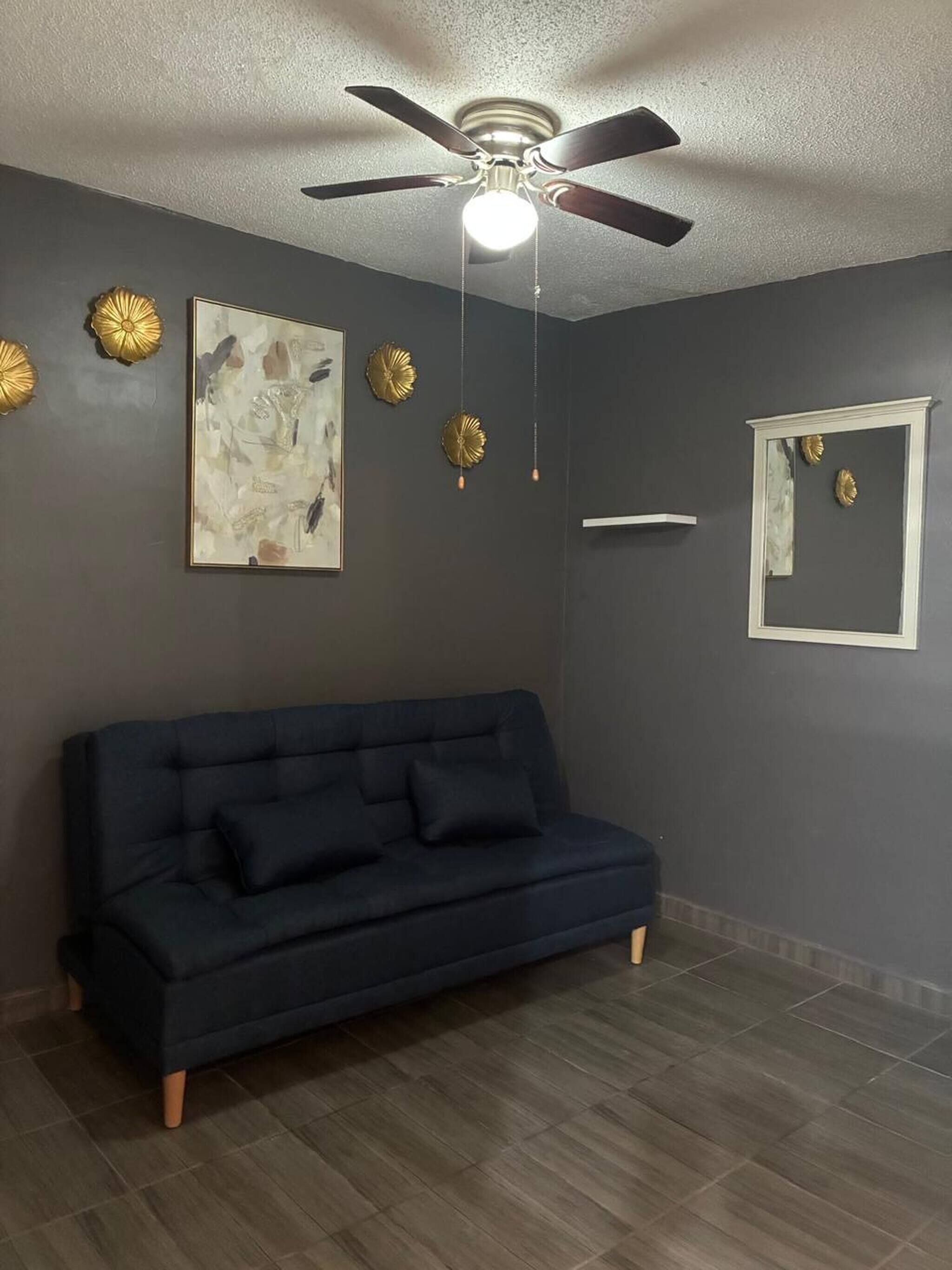 Una habitación con un sofá de color oscuro, un ventilador de techo, placas decorativas en la pared y una obra de arte enmarcada.