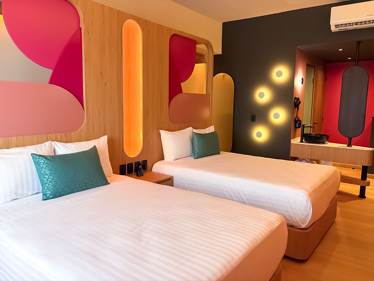 Habitación de hotel con dos camas, cabecero de madera, un espejo y diseño moderno con detalles geométricos en la pared.