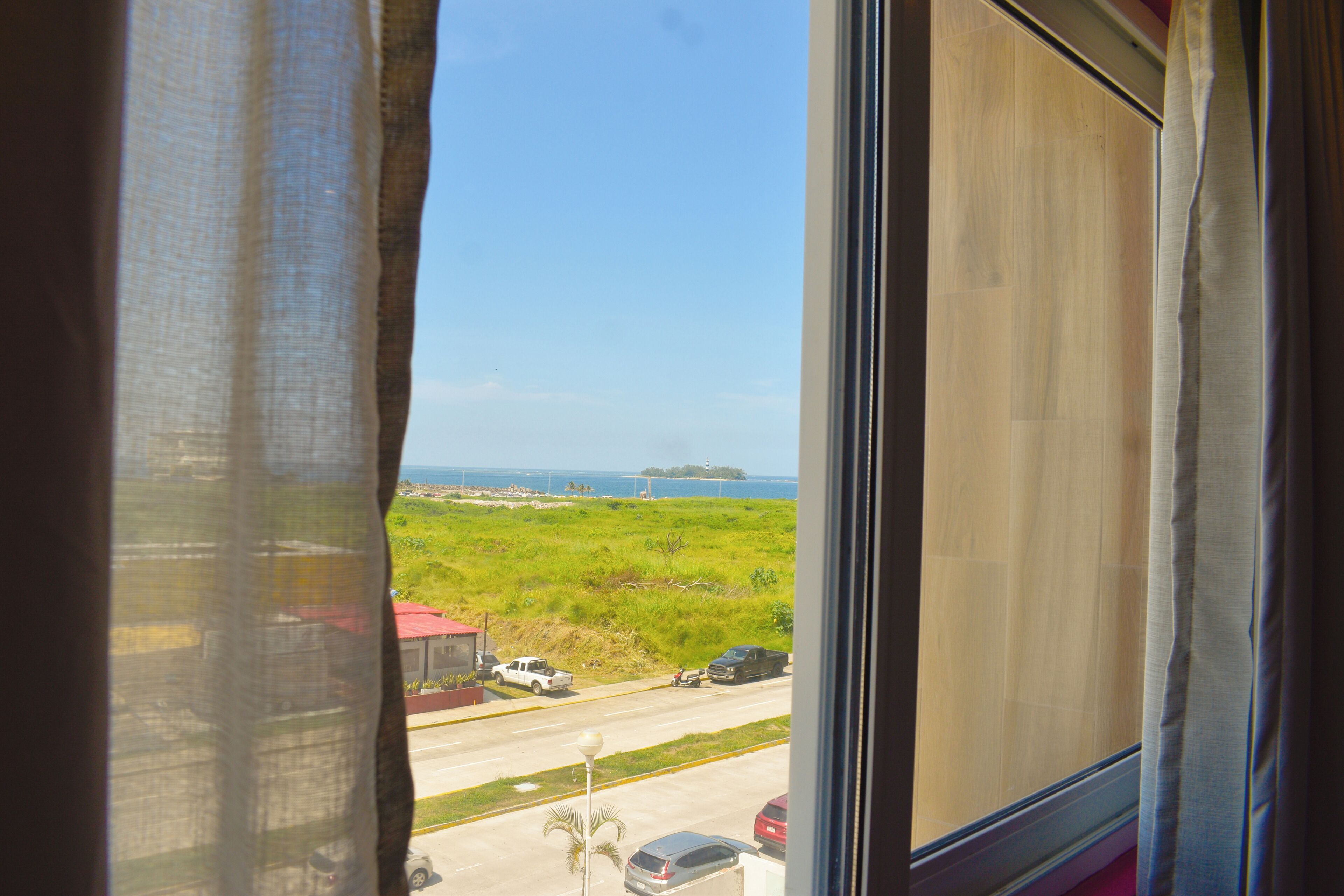 A view through a window with curtains, featuring a road, parked cars, and a distant island with a lighthouse.