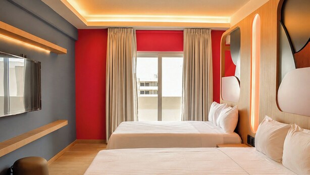 A hotel room with two beds, a large window with curtains, a flat-screen TV mounted on the wall, and a wooden floor.