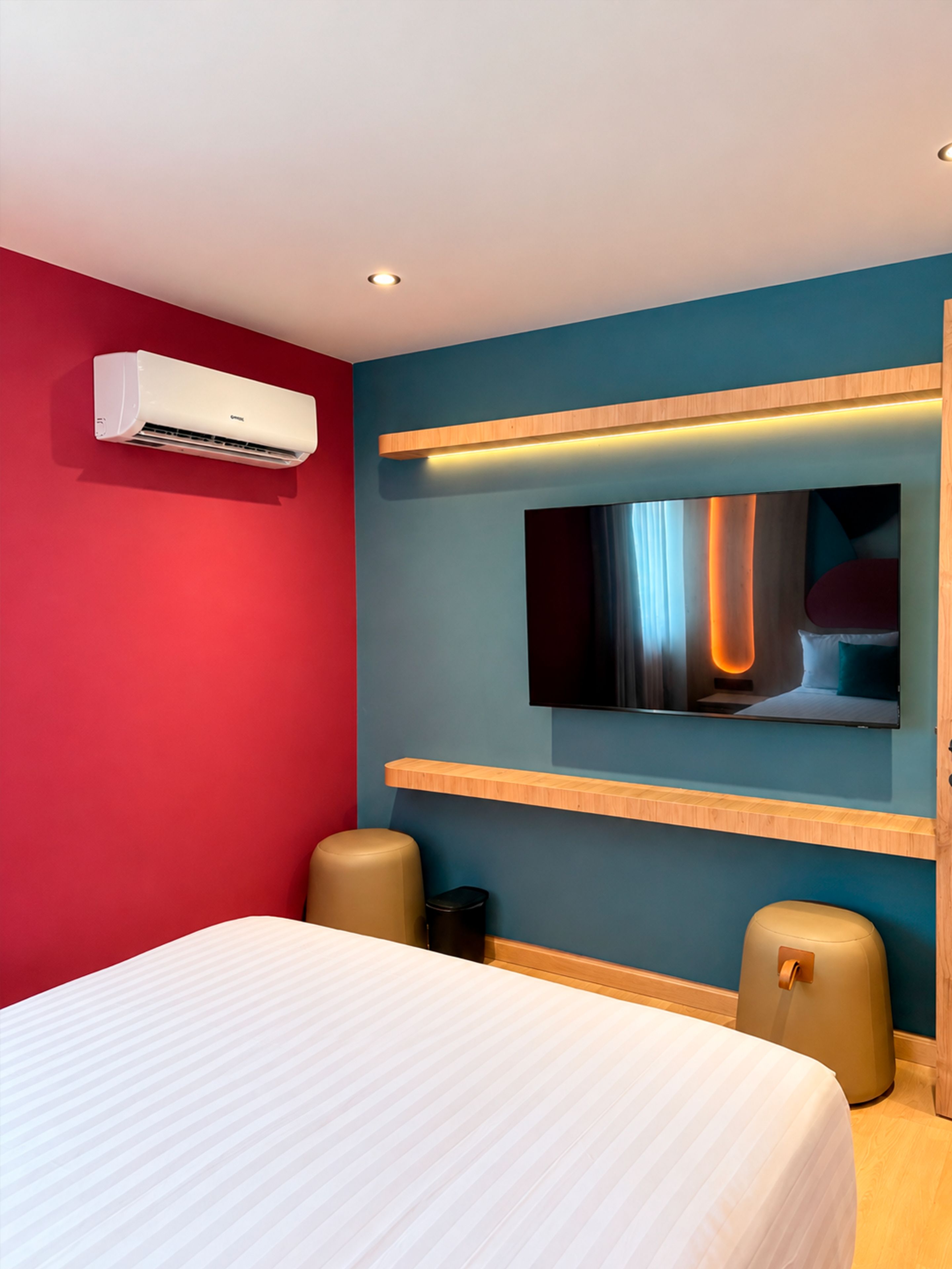 Una habitación de hotel con cama, unidad de aire acondicionado, televisor montado en la pared y dos otomanas.