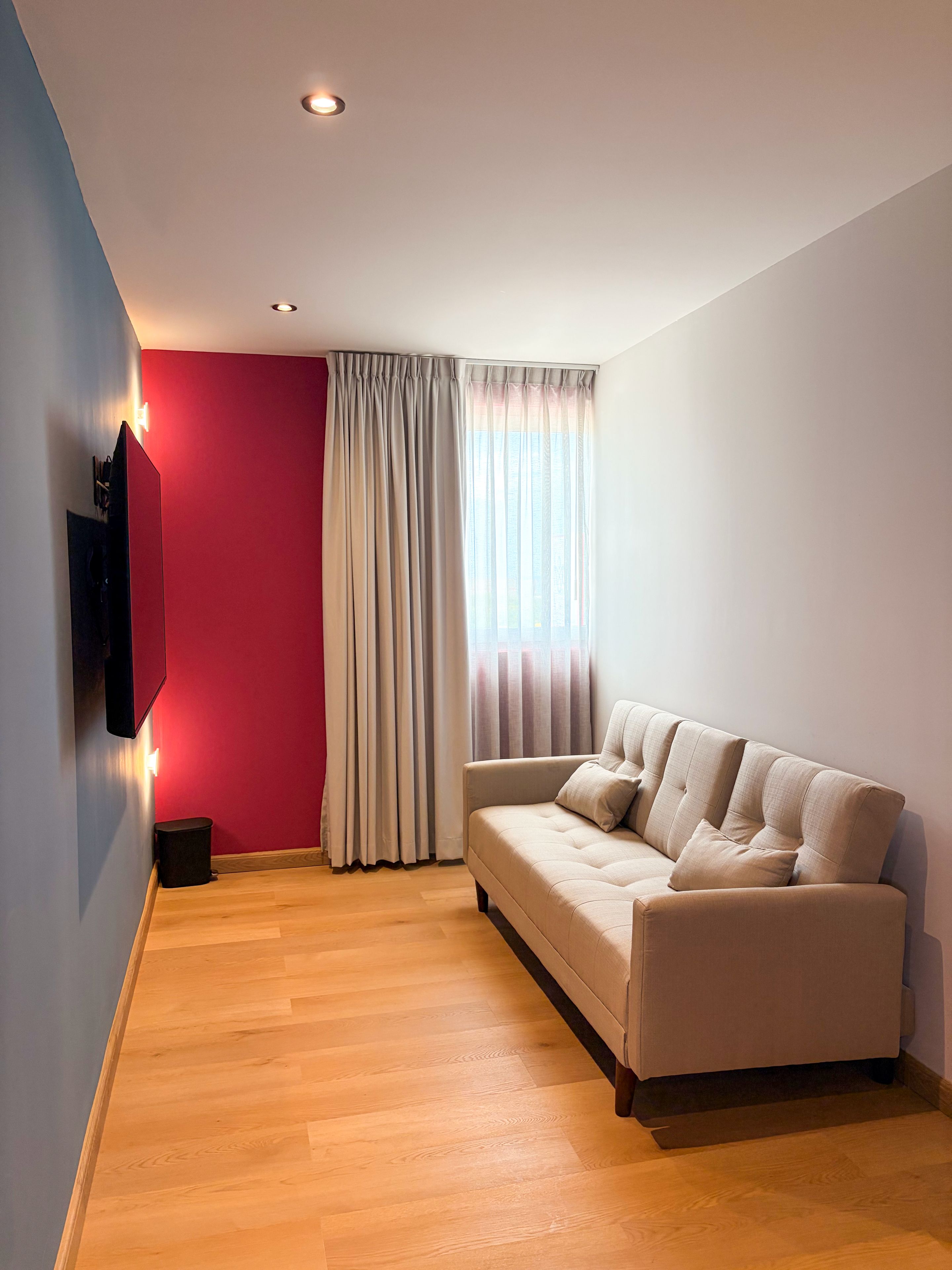Una sala de estar moderna con un sofá color beige, un televisor de pantalla plana montado en la pared y un ventanal con cortinas.
