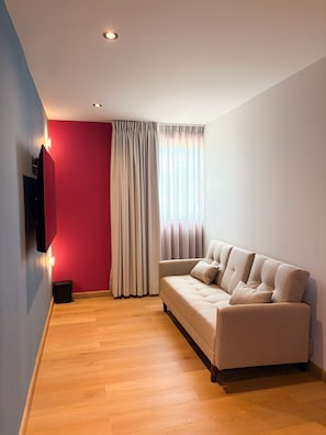 A modern living room with a beige sofa, a flat-screen TV mounted on the wall, and a large window with curtains.