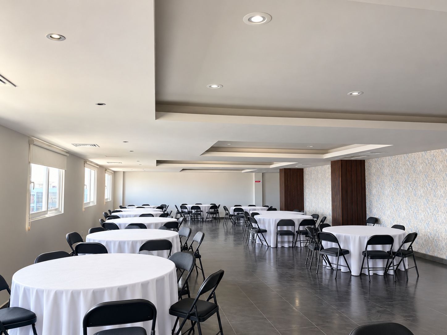 Un salón de conferencias con varias mesas redondas y sillas, luces empotradas en el techo y ventanas amplias a un lado.