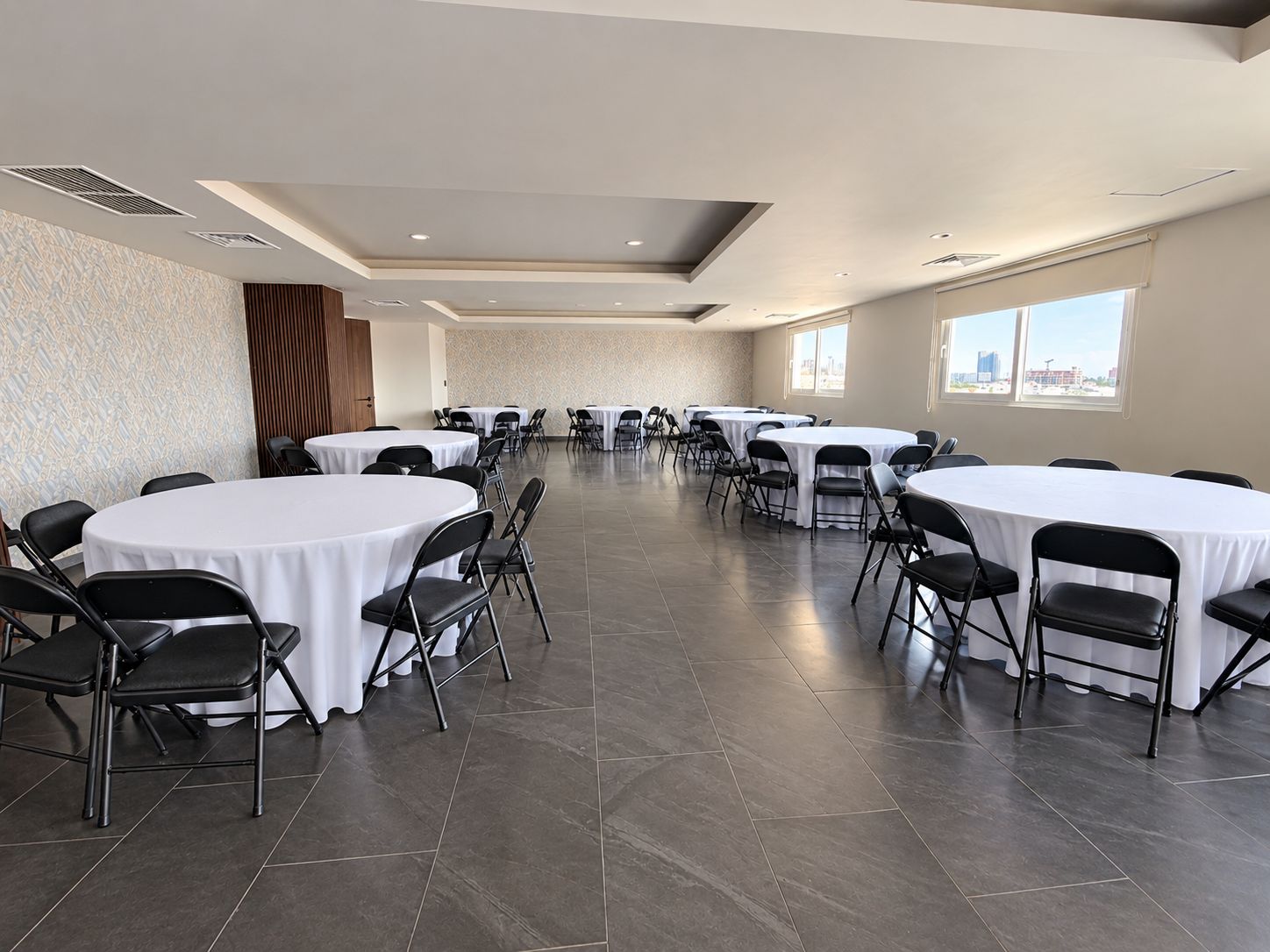 Un salón de conferencias con varias mesas redondas y sillas, ventanas amplias y un revestimiento de pared con diseño.