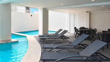 Zona de piscina interior con sillones de descanso y una mesa, rodeada de paredes y un techo con iluminación empotrada.