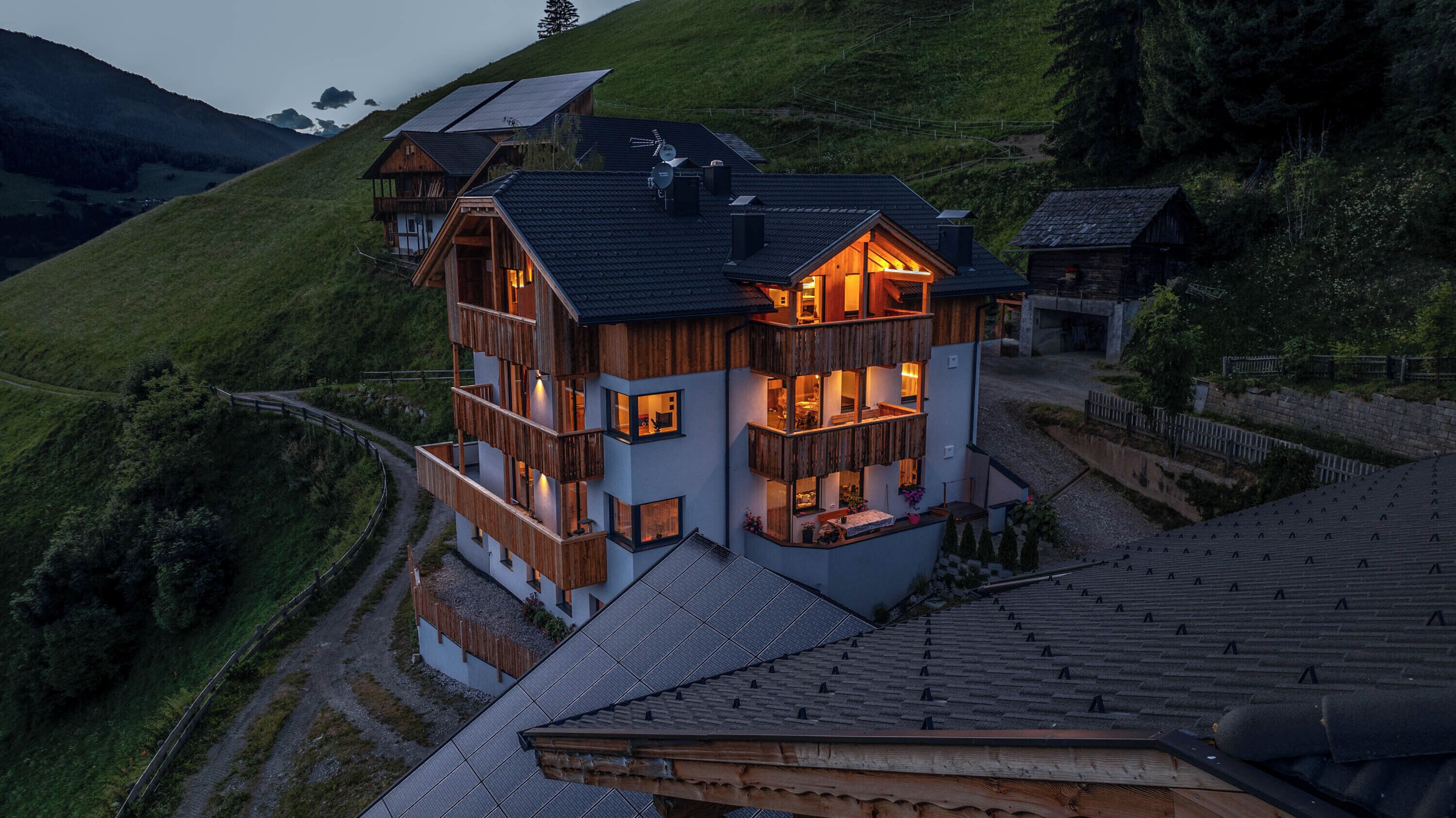 A multi-story wooden house with balconies, solar panels, and a view of a hilly landscape.