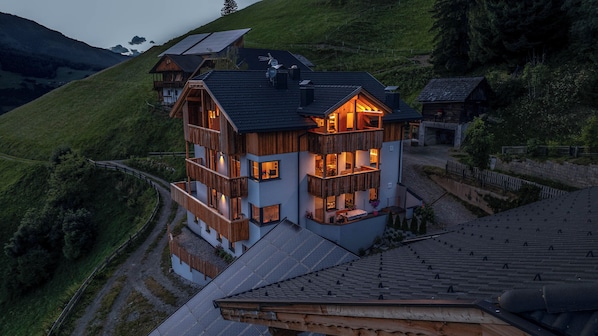 A multi-story wooden house with balconies, solar panels, and a view of a hilly landscape.