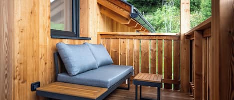 A wooden deck with a sofa and a small table, a window, and a view of greenery.