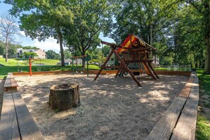 A playground with a swing set and a climbing structure in a park with sand, trees, and a picnic table. - The Fairway at the Pointe Royale (Branson)