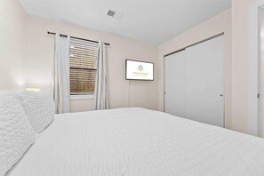 A hotel room with a bed, a window with blinds, a wall-mounted TV displaying 'GRAND WELCOME,' and a door. - The Fairway at the Pointe Royale (Branson)