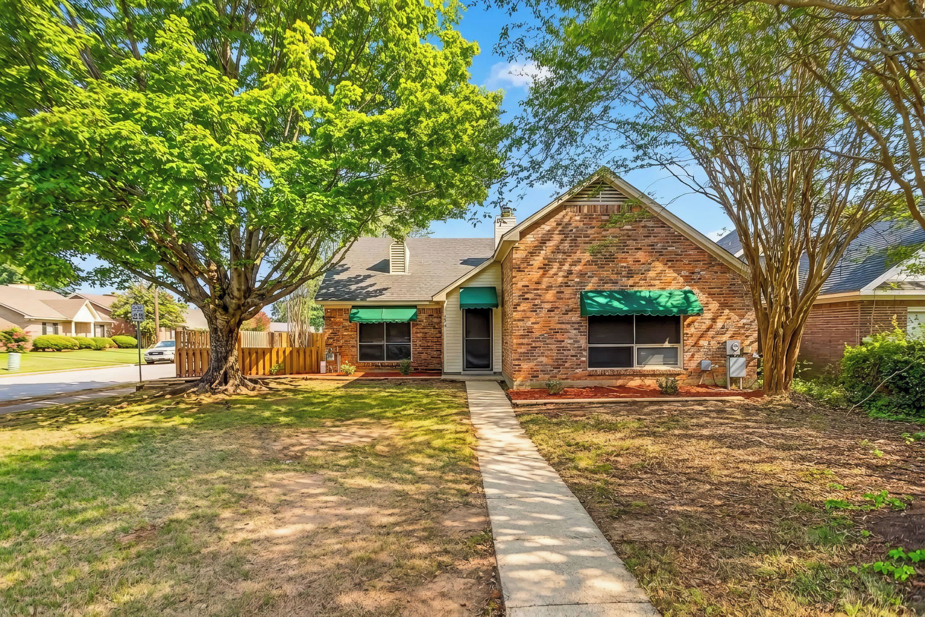 A brick house with a green awning, a front yard with a tree, and a sidewalk leading to the entrance.