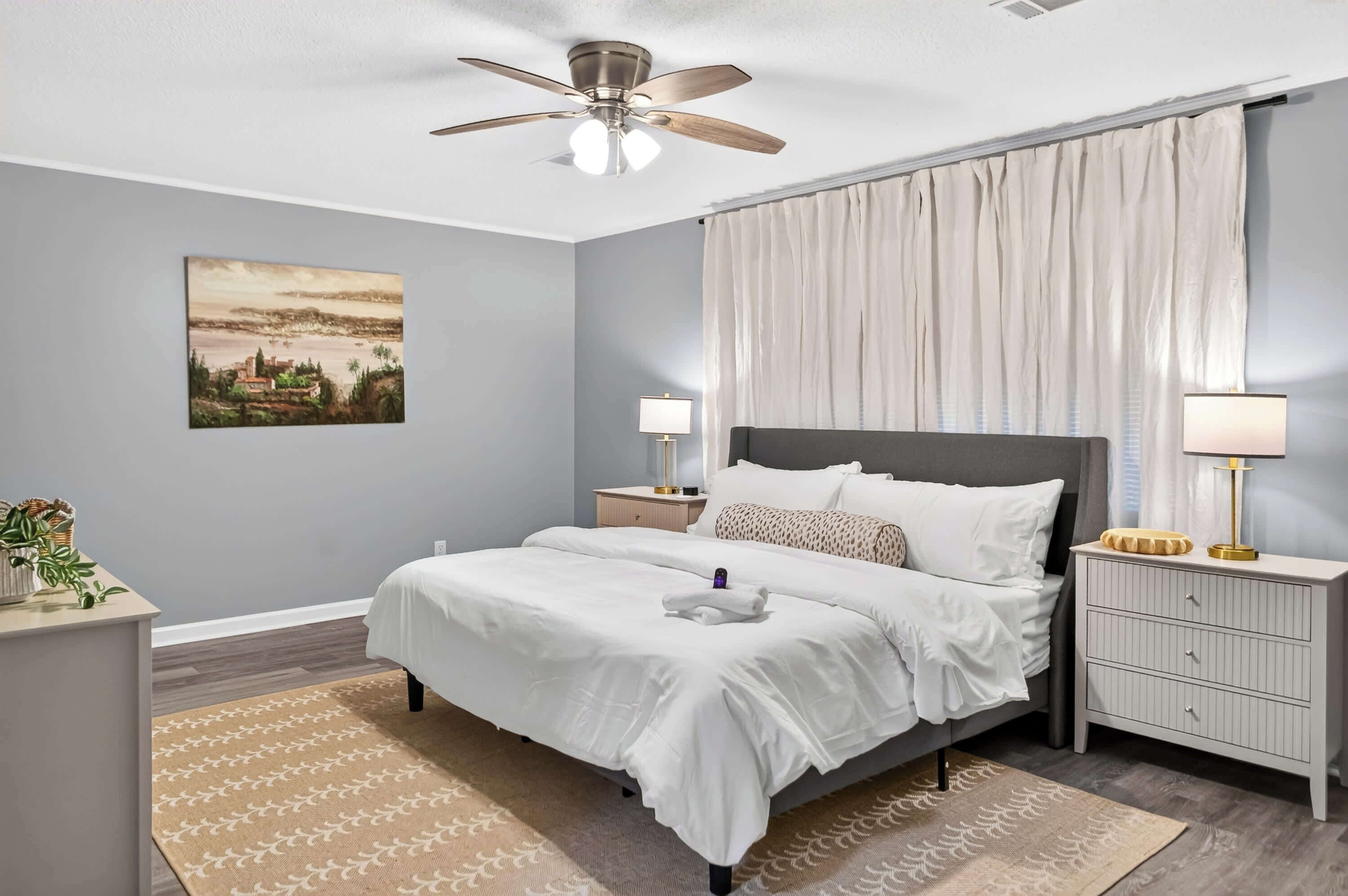 A bedroom with a bed, nightstands, a ceiling fan, a painting, and a window with curtains.