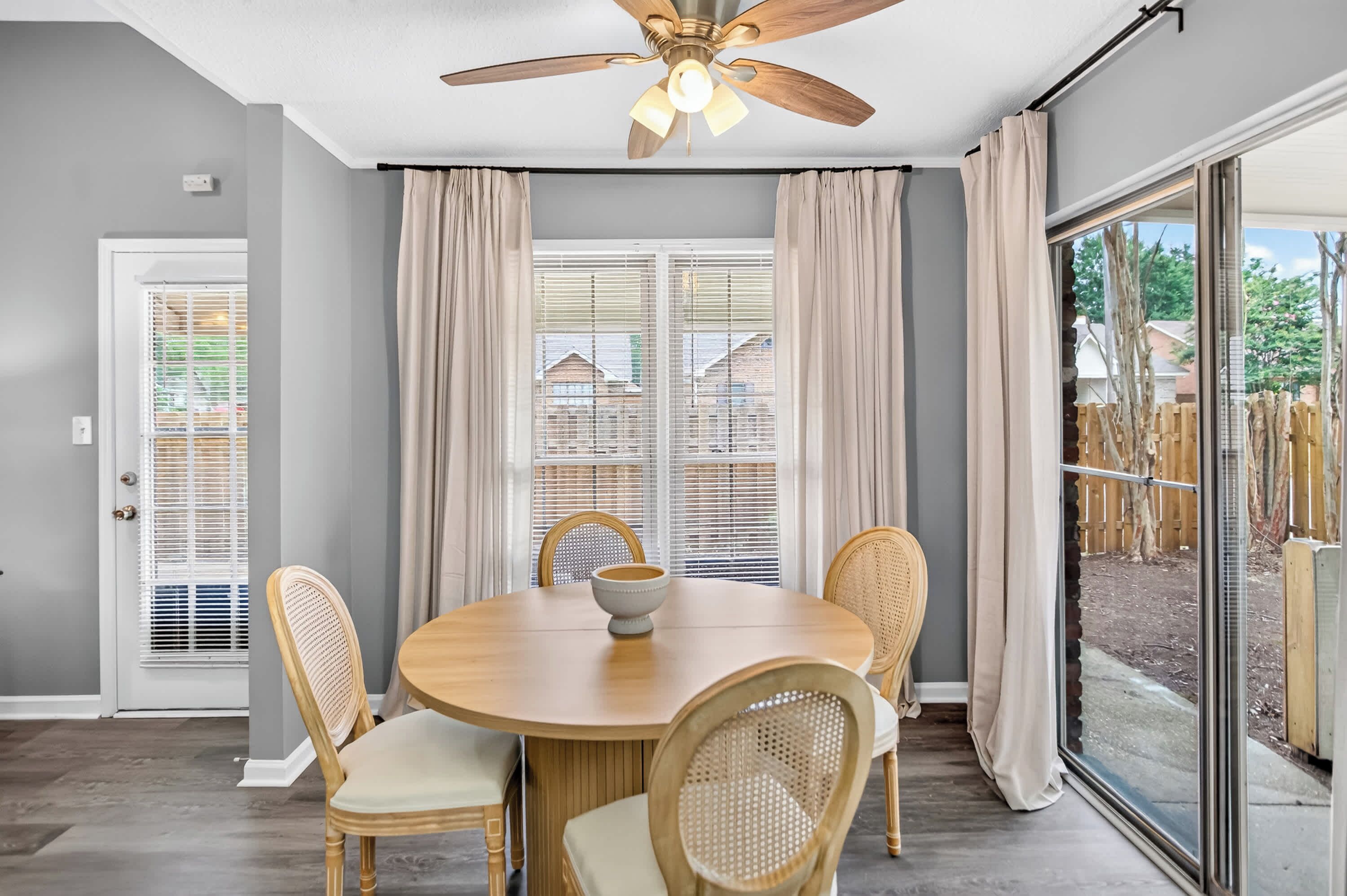 Un coin repas comprenant une table ronde, quatre chaises, un ventilateur de plafond et de grandes fenêtres avec des rideaux.