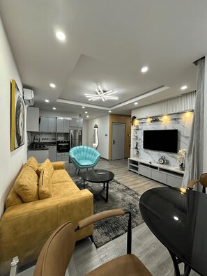 A modern living room with a sofa, a teal chair, a dining table, and a TV mounted on a marble wall. - Premium 1-Bedroom Apartment in Lekki 1 with Fast Wi-Fi & 24/7 Power (Lekki)
