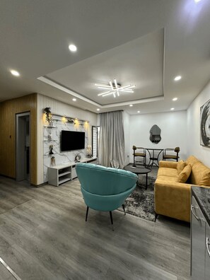 A modern living room with a teal armchair, a yellow sofa, a TV mounted on a marble wall, and a dining area with a table and chairs. - Premium 1-Bedroom Apartment in Lekki 1 with Fast Wi-Fi & 24/7 Power (Lekki)