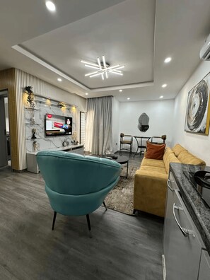 A modern living room with a teal armchair, a yellow sofa, a dining area with a table and chairs, a TV mounted on the wall, and a contemporary ceiling light fixture. - Premium 1-Bedroom Apartment in Lekki 1 with Fast Wi-Fi & 24/7 Power (Lekki)