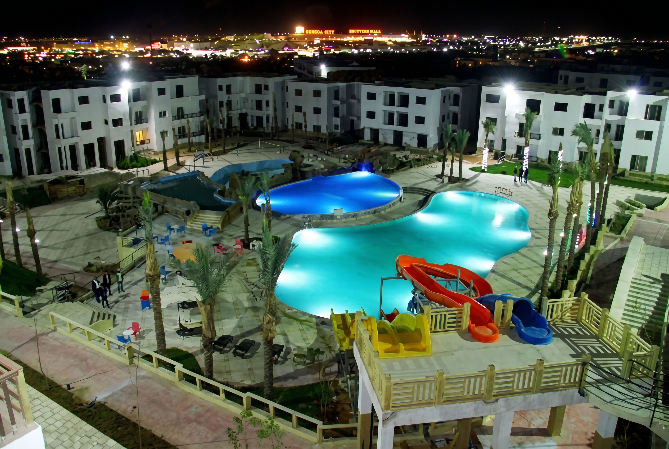 Un complexe hôtelier doté de plusieurs piscines, d’un toboggan et offrant une vue sur les lumières d’une ville lointaine la nuit.