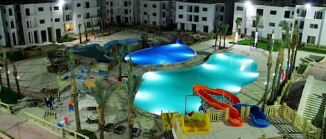 Un complexe hôtelier doté de plusieurs piscines, d’un toboggan et offrant une vue sur les lumières d’une ville lointaine la nuit.