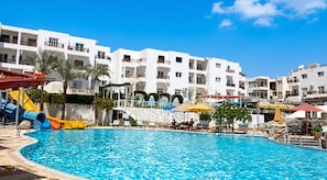 A swimming pool with a water slide, lounge chairs, umbrellas, and a building with balconies in the background.