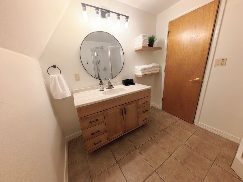 A bathroom with a wooden vanity, a round mirror, a towel rack, and a door.