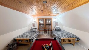 A room with a pool table, two beds with checkered bedding, a wooden ceiling, and a window with blinds.