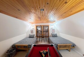 A room with a pool table, two beds with checkered bedding, a wooden ceiling, and a window with blinds.