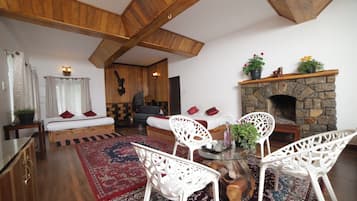 A cozy living room with a stone fireplace, wooden beams, a sofa, and white chairs.