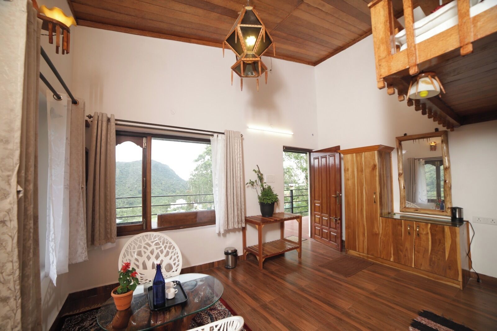 A spacious room with a wooden ceiling, a large window with a view of greenery, a wooden cabinet, and a glass table with a tray.