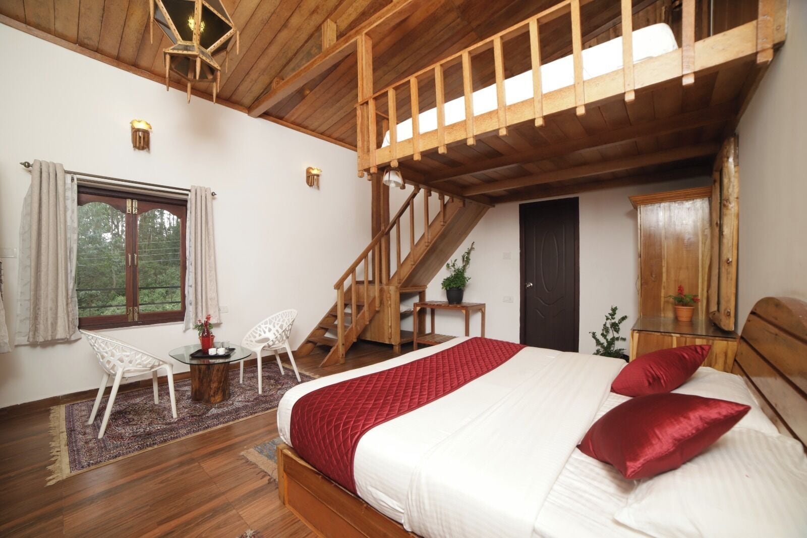 A bedroom with a wooden ceiling, a bed with red pillows, a window with curtains, a white chair, and a small table.