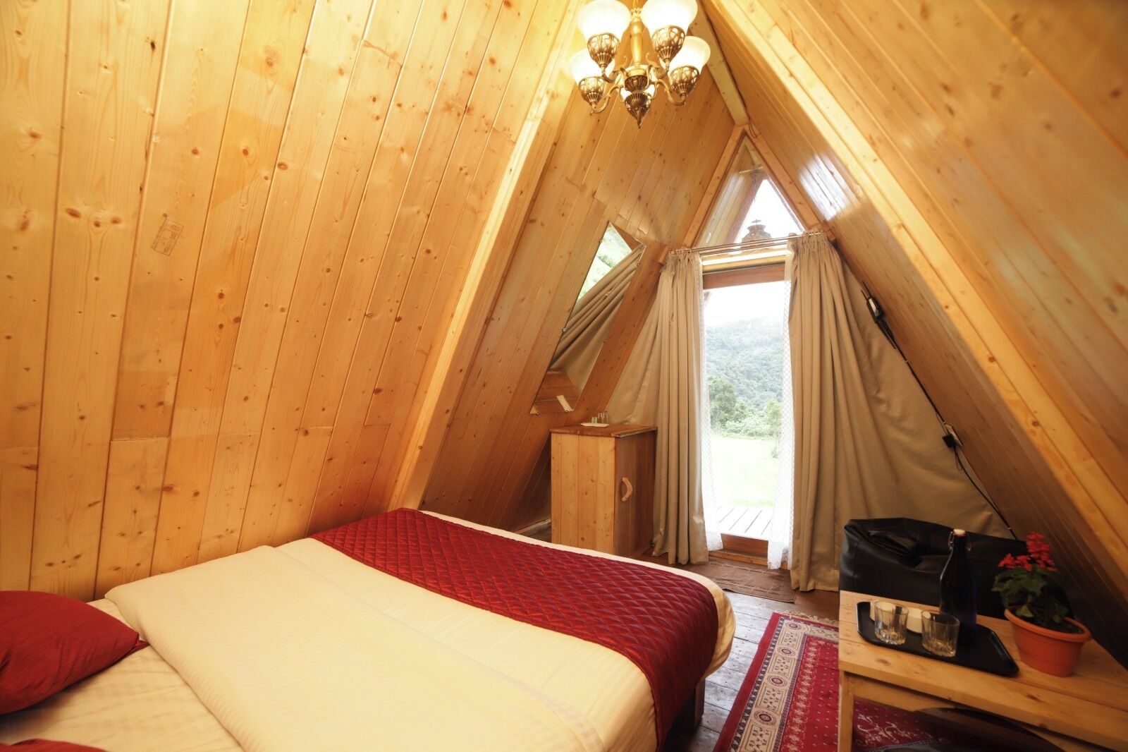 Una habitación acogedora, con techo de madera, una cama, una pequeña mesa con una planta y una ventana con cortinas.