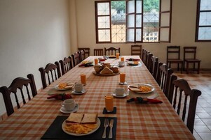 Una larga mesa de comedor con sillas, preparada para el desayuno, con platos de comida, tazas y cubiertos.