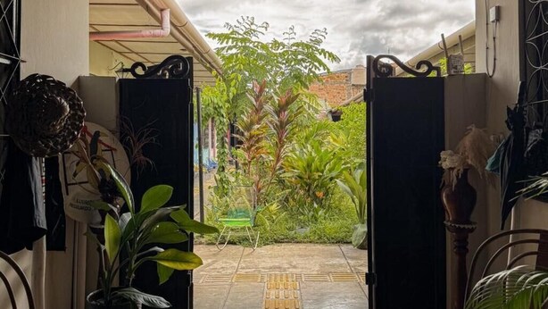 A covered outdoor area with a wooden floor leading to a garden. There are potted plants and a chair.